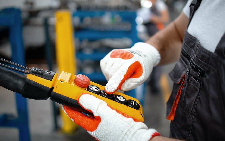 Saubere Kranflasche Das Bild zeigt die Hände eines Mitarbeiters in einer Arbeitshalle. Er trägt Handschuhe und hält eine Kranflasche in der linken Hand. Mit dem rechten Zeigefinger drückt der Mitarbeiter auf einen Knopf auf der Kranflasche. Link zum Artikel.