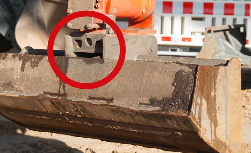 Das Bild zeigt ein angekoppeltes Anbaugerät einer Erdbaumaschine, das zwar in der Aufnahmeklaue liegt, aber nicht verriegelt ist. Ein roter Kreis zeigt genau die Stelle an. In diesem Fall besteht Absturzgefahr beim Hochheben und Schwenken des Anbaugerätes. 