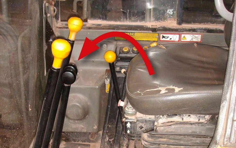 Das Bild zeigt den Blick in die Fahrerkabine eines Minibaggers. Ein roter Pfeil verdeutlicht, wo der Sitz umgeklappt wurde, um an die Batterie heranzukommen. Dieser drückte dann gegen die Fahrhebel. Dadurch kam es zu einer unerwarteten Bewegung des Minibaggers und zum Unfall.