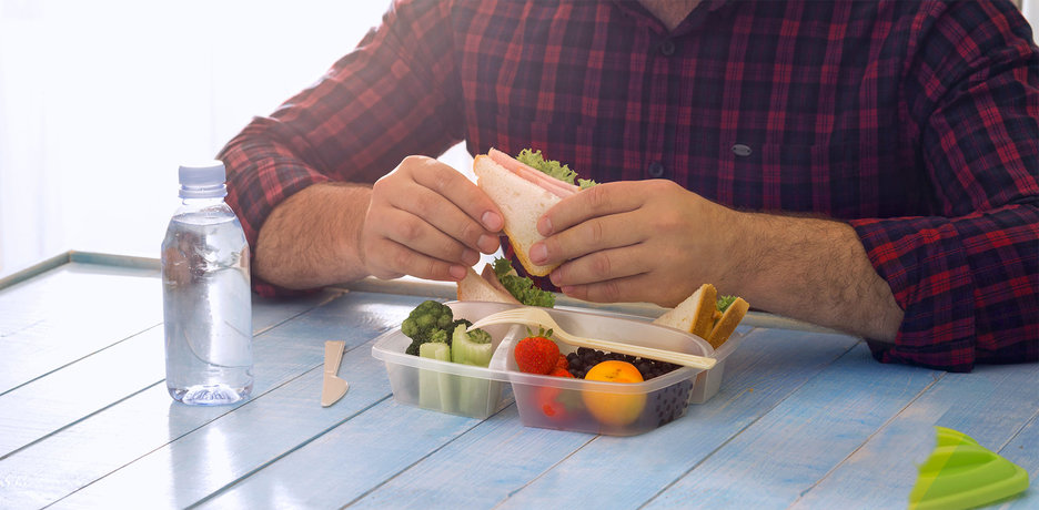 Zu sehen ist der Oberkörper eines  Mannes, der am Tisch sitzt und isst. Vor ihm steht eine geöffnete Brotdose mit gesundem Obst und Gemüse. In den Händen hält er ein belegtes Sandwich. Auf der linken Seite des Bildes steht eine mit Wasser gefüllte Plastikflasche. Link zur vergrößerten Darstellung des Bildes.
