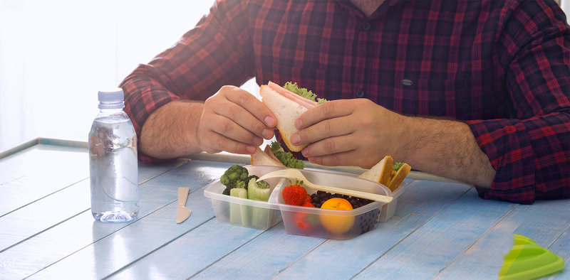 Zu sehen ist der Oberkörper eines  Mannes, der am Tisch sitzt und isst. Vor ihm steht eine geöffnete Brotdose mit gesundem Obst und Gemüse. In den Händen hält er ein belegtes Sandwich. Auf der linken Seite des Bildes steht eine mit Wasser gefüllte Plastikflasche.