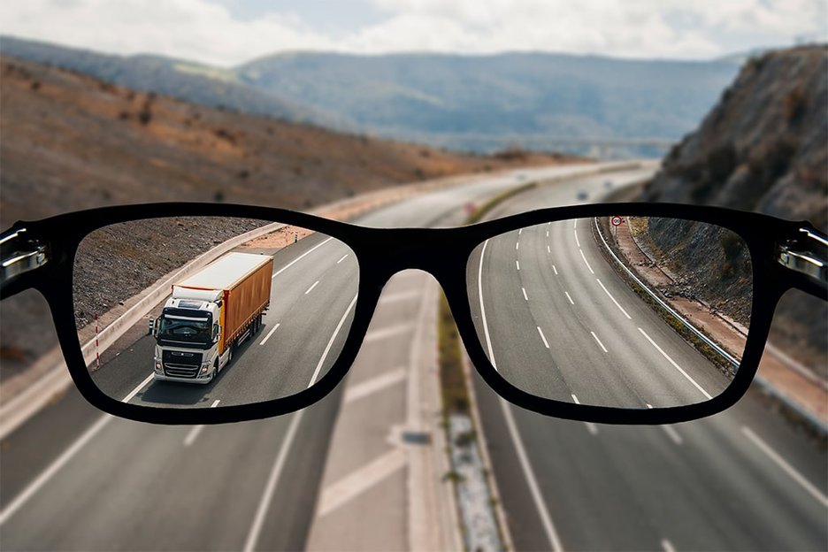 Zu sehen ist eine Autobahn mit mehreren Fahrspuren. Davor befindet sich eine schwarze Brille. Der Blick durch die Brillengläser zeigt vergrößerte Fahrspuren und einen Lkw auf der Autobahn. Dies verdeutlicht die bessere Sehleistung mit Brille im Verkehr. Link zur vergrößerten Darstellung des Bildes.