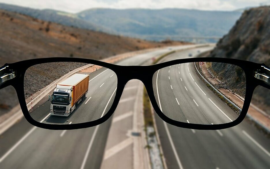 Zu sehen ist eine Autobahn mit mehreren Fahrspuren. Davor befindet sich eine schwarze Brille. Der Blick durch die Brillengläser zeigt vergrößerte Fahrspuren und einen Lkw auf der Autobahn. Dies verdeutlicht die bessere Sehleistung mit Brille im Verkehr.
