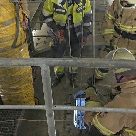 Das Bild zeigt den Blick auf das Mannloch der Kugelmühle. Darum stehen Feuerwehrleute und Mitarbeiter des Betriebs. Links hält eine Person eine Rettungstrage, die aussieht, wie ein großer gelber Plastiksack. In das Loch führen 2 Seile einer Seilwinde. Link zur vergrößerten Darstellung des Bildes.