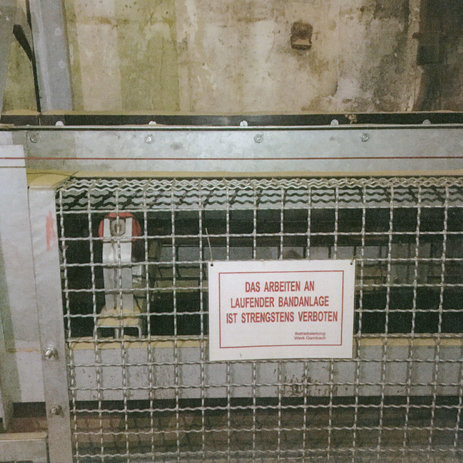 Das Bild zeigt ein Warnschild an einem Förderband, das die Arbeit an laufender Bandanlage strengstens verbietet. Link zur vergrößerten Darstellung des Bildes.