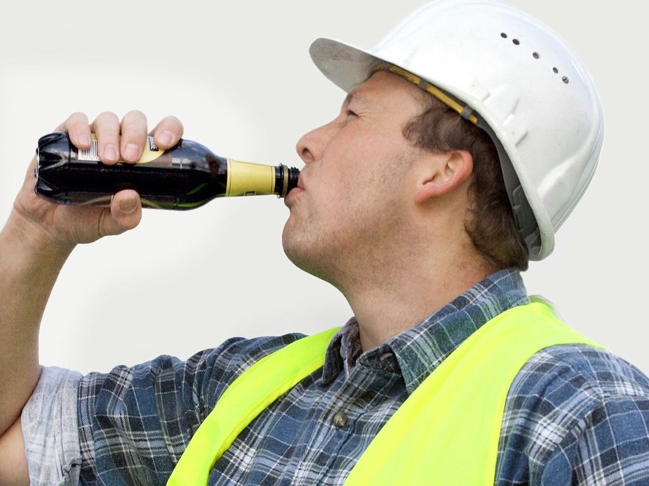 Ein Mitarbeiter mit Schutzhelm und Warnweste hat den rechten Arm gehoben und trinkt aus einer Bierflasche am Arbeitsplatz. Link zur vergrößerten Darstellung des Bildes.