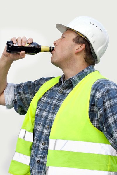Ein Mitarbeiter mit Schutzhelm und Warnweste hat den rechten Arm gehoben und trinkt aus einer Bierflasche am Arbeitsplatz.