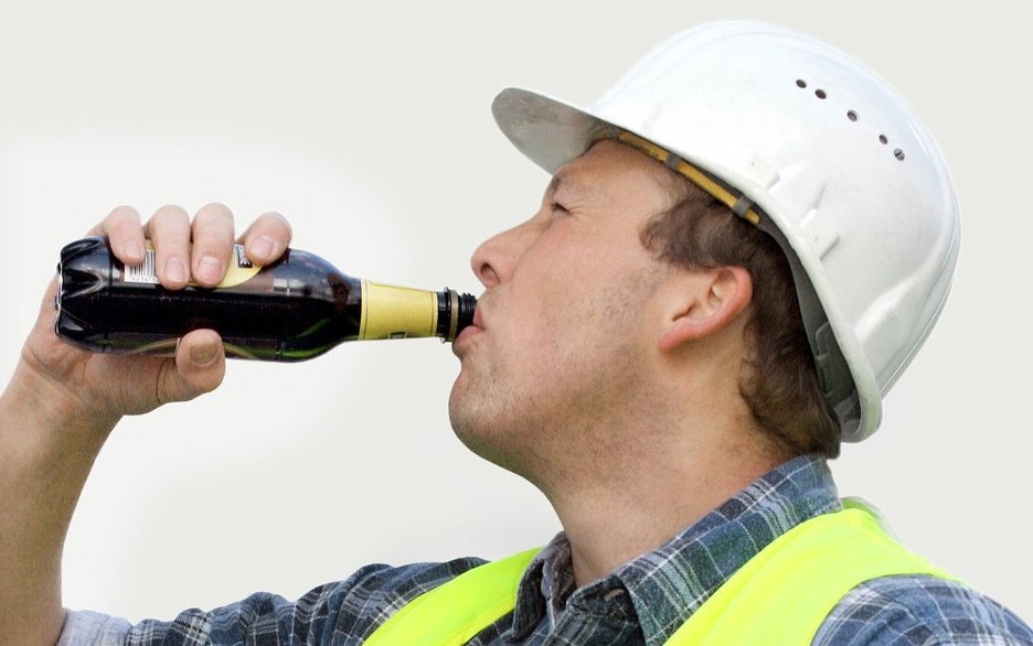 Ein Mitarbeiter mit Schutzhelm und Warnweste hat den rechten Arm gehoben und trinkt aus einer Bierflasche am Arbeitsplatz. Link zum Artikel.