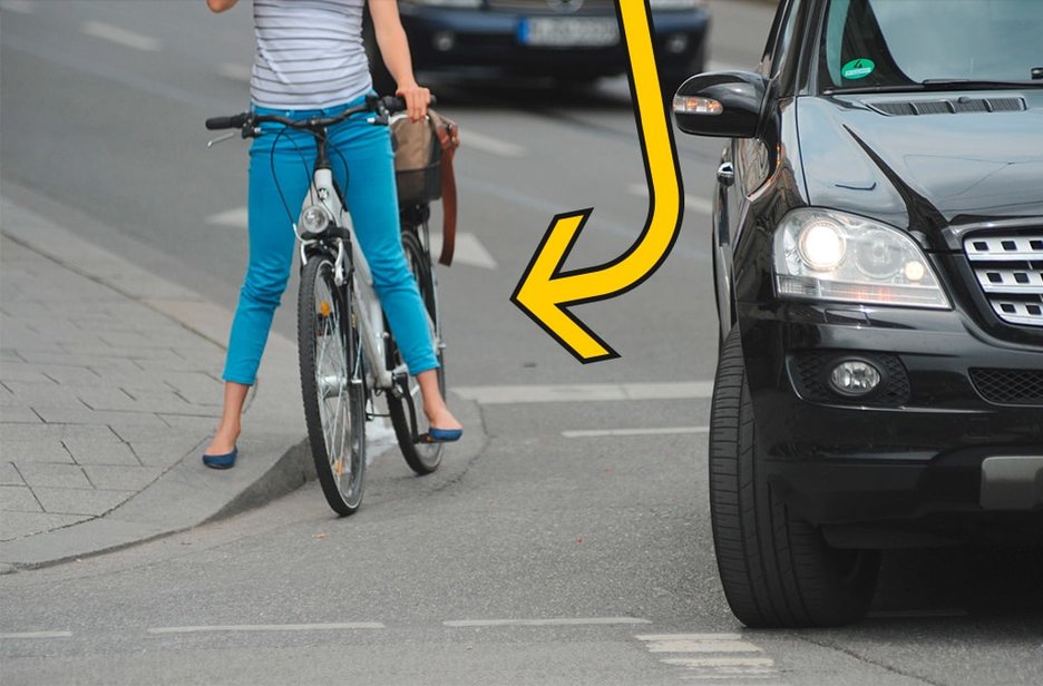 An einer Kreuzung befindet sich ein rechtsabbiegender Pkw. Rechts daneben wartet eine Radfahrerin auf der Straße ab, was passiert.