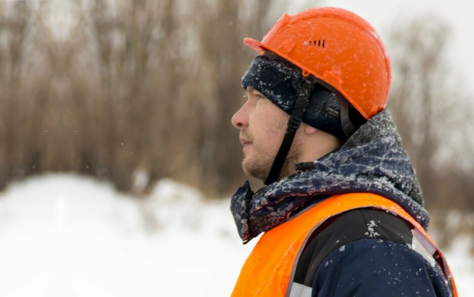 Das Foto zeigt einen Mitarbeiter im Portrait von der linken Seite im Winter, der unter seinem Schutzhelm eine Mütze trägt und Winterschutzkleidung mit Warnweste. Er schaut direkt links aus dem Bild heraus.