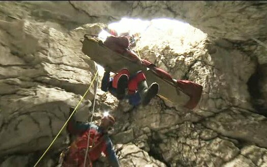 Das Bild zeigt, die spektakuläre Rettung des Höhlenforschers Johann Westhauser angeseilt auf einer Trage aus der Riesending-Höhle. Zwei Retter schweben angeseilt neben der Trage. Der Blick ist von unten aus Höhle hinaus ans Tageslicht.
