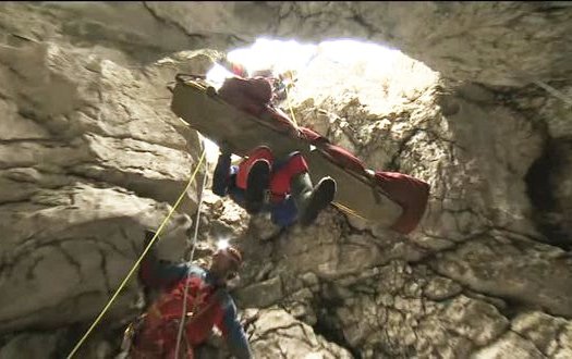 Das Bild zeigt, die spektakuläre Rettung des Höhlenforschers Johann Westhauser angeseilt auf einer Trage aus der Riesending-Höhle. Zwei Retter schweben angeseilt neben der Trage. Der Blick ist von unten aus Höhle hinaus ans Tageslicht. Link zur vergrößerten Darstellung des Bildes.