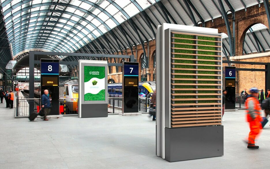 Das Bild zeigt zwei CityTrees in einem Bahnhofsgebäude in Form von Stelen. Auf der einen Seite der CityTrees befindet sich eine grüne, luftreinigende Mooswand, auf der anderen ein 75-Zoll-LCD-Bildschirm.  Link zum Artikel.
