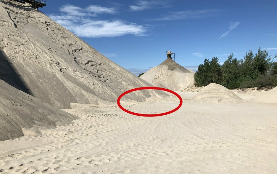 Zu sehen ist der Blick auf eine Halde. In der Mitte ist ein roter Kreis. Dies ist der Ort, wo der Unfall passierte. Dabei stieß ein Radlader, der Sand verkippte, mit einem Fußgänger zusammen. Die leblose Person wurde später an der Vorratshalde im Sand gefunden. Link zum Artikel.