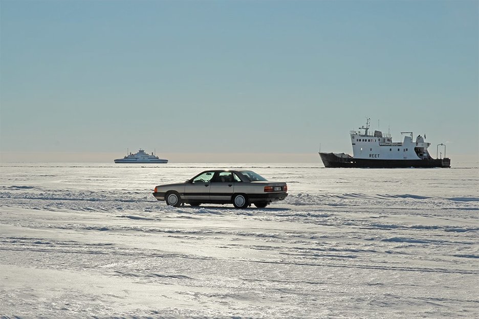 Zu sehen ist ein Pkw, der auf der zugefrorenen Eisfläche der Ostsee zur Insel Vormsi in Estland fährt. Im Hintergrund sind Schiffe zu sehen, die auf dem offenen Teil der Ostsee fahren. Link zur vergrößerten Darstellung des Bildes.