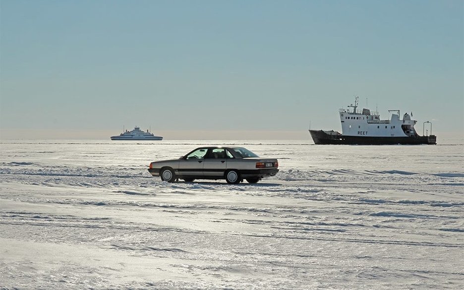 Zu sehen ist ein Pkw, der auf der zugefrorenen Eisfläche der Ostsee zur Insel Vormsi in Estland fährt. Im Hintergrund sind Schiffe zu sehen, die auf dem offenen Teil der Ostsee fahren.