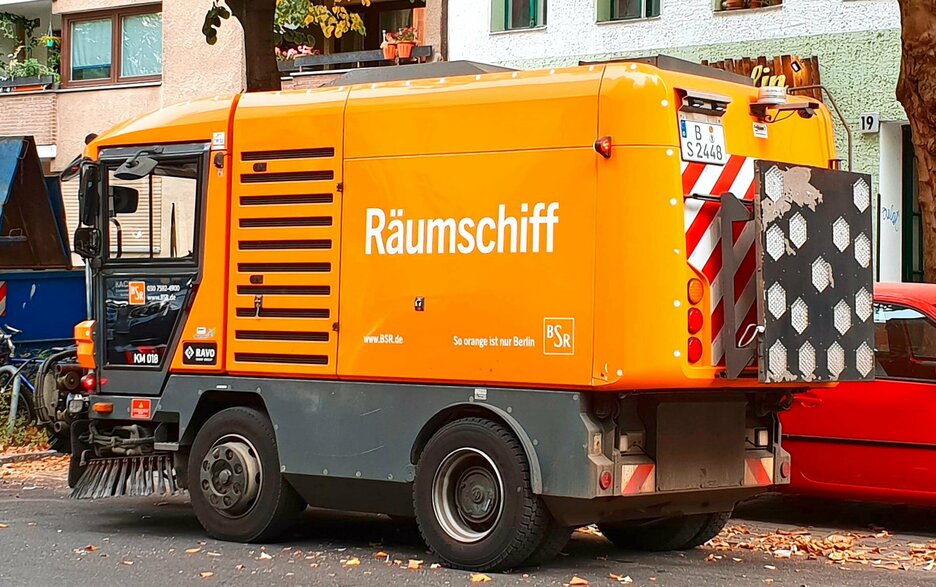 Das Bild zeigt eine moderne, orangefarbene Kehrmaschine der Berliner Stadtreinigung. Auf der Seite steht der witzige Name „Räumschiff“.  