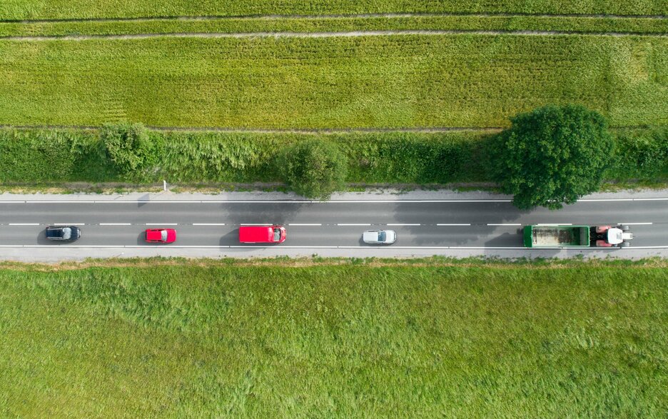 Das Foto zeigt eine Verkehrssituation auf einer schnurgeraden Landstraße aus der Vogelperspektive. Zu sehen sind drei Autos und ein Transporter, die hinter einem Trecker mit leerem Anhänger herfahren. Hier ist höchste Vorsicht beim Überholen geboten! Die Straße ist baumbestanden und führt durch Wiesen und landwirtschaftliche Felder.  Link zum Artikel.