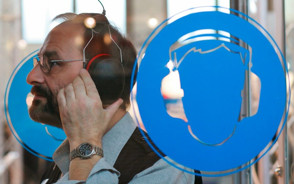 Zu sehen ist ein Mann mit Kopfhörern, der sich in einer Kabine mit Gehörschutzsymbolen Hörbeispiele anhört Link zum Artikel.