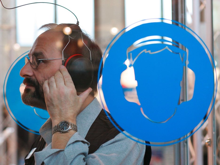 Zu sehen ist ein Mann mit Kopfhörern, der sich in einer Kabine mit Gehörschutzsymbolen Hörbeispiele anhört. Link zur vergrößerten Darstellung des Bildes.