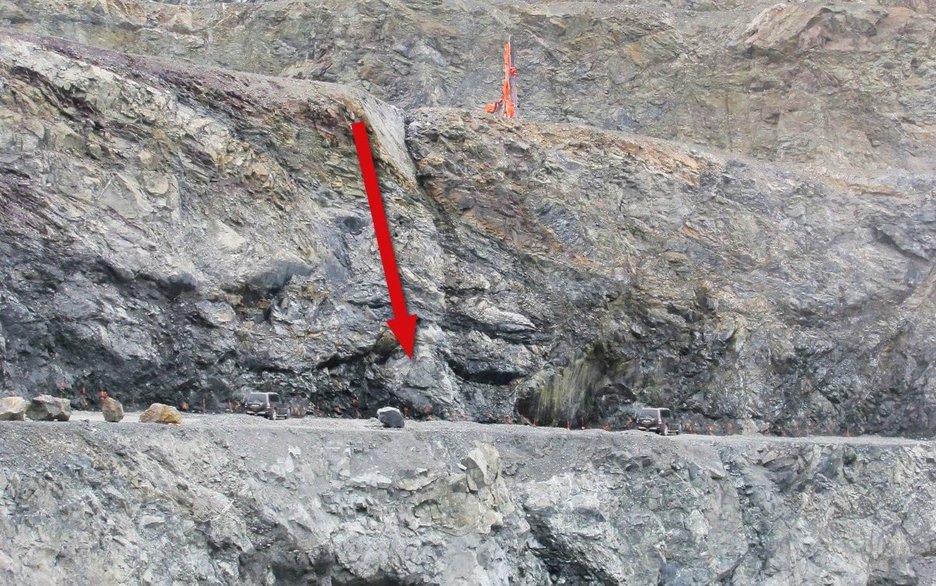 Das Bild zeigt die Bruchwand in einem Steinbruch. Auf der oberen Sohle ist ein Bohrgerät zu sehen, das nah an der Abbruchkante steht. Ein roter Pfeil links daneben führt entlang einer Rinne abwärts auf die untere Sohle. Hier stürzte ein Bohrmaschinist 25 m in die Tiefe.