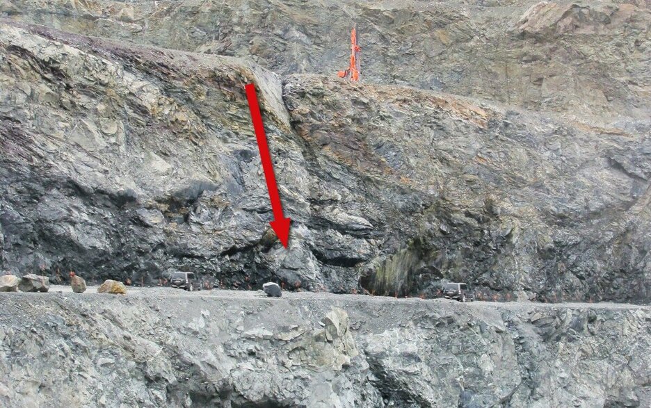 Das Bild zeigt die Bruchwand in einem Steinbruch. Auf der oberen Sohle ist ein Bohrgerät zu sehen, das nah an der Abbruchkante steht. Ein roter Pfeil links daneben führt entlang einer Rinne abwärts auf die untere Sohle. Hier stürzte ein Bohrmaschinist 25 m in die Tiefe. Link zum Artikel.