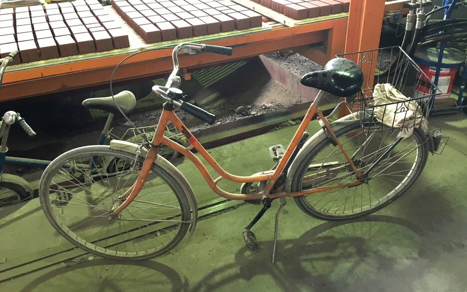 Das Bild zeigt zwei in die Jahre gekommene Fahrräder von der Seite in einer Werkhalle. Eines ist orange und steht im Vordergrund, das andere ist dunkel und steht dahinter, etwas unterhalb in einer Rille. Beide Fahrräder haben kein Licht, keine Reflektoren und Rückstrahler. Sie sind nicht verkehrssicher. Link zum Artikel.