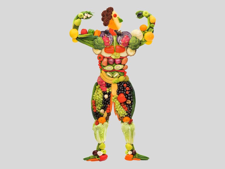 Auf dem Foto ist ein aus Obst und Gemüse geformter Mann zu sehen, der muskulös wirkt und vor Kraft strotzend beide Arme erhoben hat, um seine Muskeln zu zeigen.