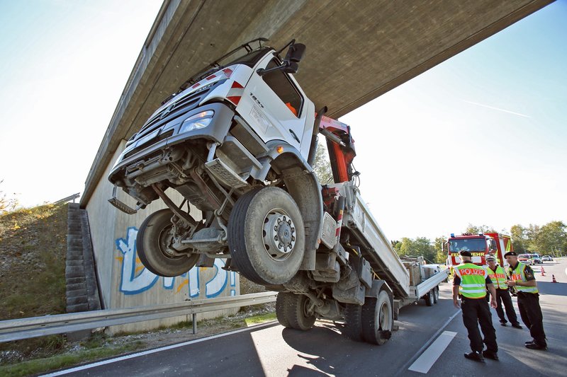 Das Bild zeigt die Vorderansicht eines Unfalls, bei dem ein Lkw mit seinem nicht ganz richtig eingefahrenen Kranausleger unter einer Brücke hängengeblieben ist. Der Lkw ist dabei vorn steil in die Höhe gereckt und steht nur noch auf den Hinterrädern.