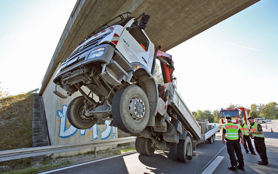 Das Bild zeigt die Vorderansicht eines Unfalls, bei dem ein Lkw mit seinem nicht ganz richtig eingefahrenen Kranausleger unter einer Brücke hängengeblieben ist. Der Lkw ist dabei vorn steil in die Höhe gereckt und steht nur noch auf den Hinterrädern.