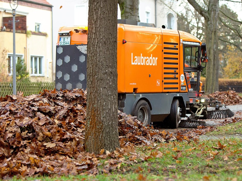 Das Bild zeigt eine moderne, orangefarbene Kehrmaschine der Berliner Stadtreinigung, die große Mengen Laub von der Straße entfernt. Auf der Seite steht der witzige Name „Laubrador“. 