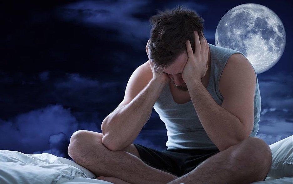 Ein junger Mann sitzt nachts schlaflos im Schneidersitz in seinem Bett. Er hat die Ellenbogen auf seine Knie gestützt und hält seinen Kopf mit den Händen fest. Hinter ihm rechts über seiner Schulter ist ein großer Mond am Nachthimmel zu sehen. Link zum Artikel.