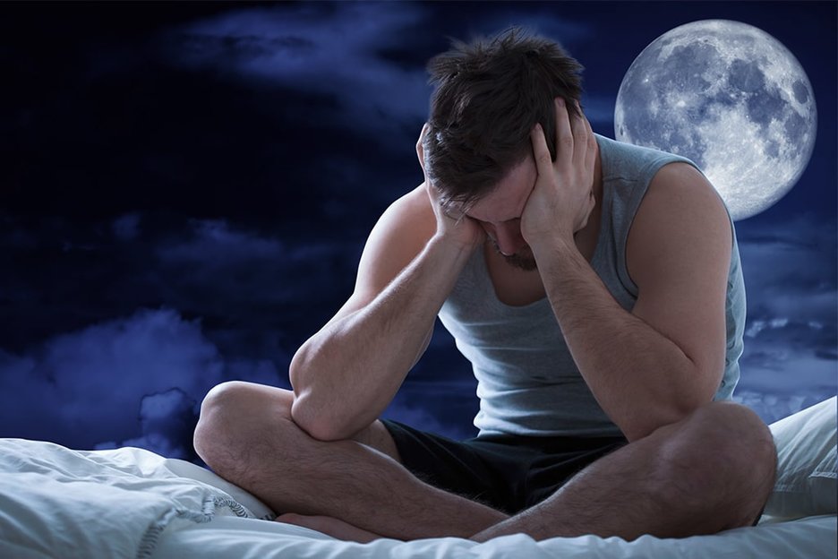 Ein junger Mann sitzt nachts schlaflos im Schneidersitz in seinem Bett. Er hat die Ellenbogen auf seine Knie gestützt und hält seinen Kopf mit den Händen fest. Hinter ihm rechts über seiner Schulter ist ein großer Mond am Nachthimmel zu sehen. Link zur vergrößerten Darstellung des Bildes.