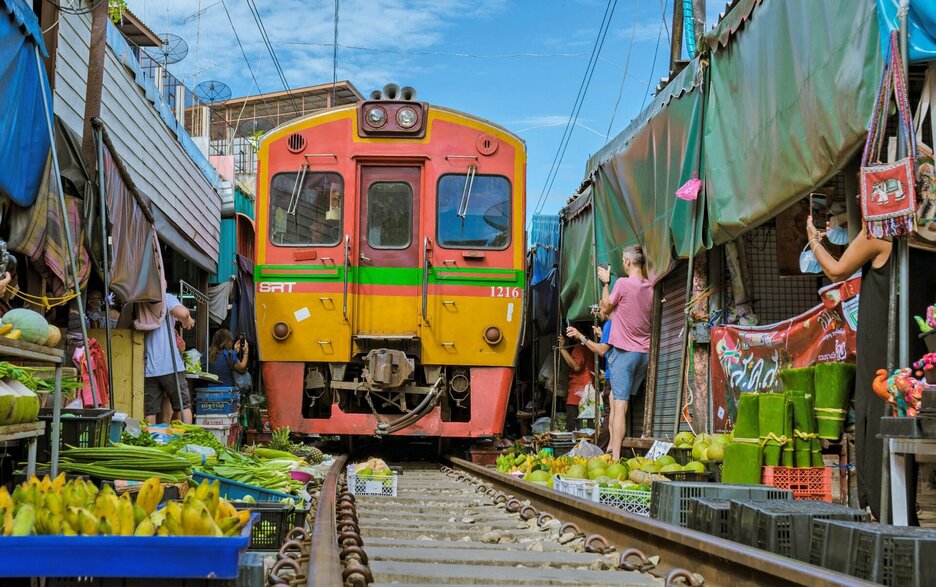Das Bild zeigt den Mae-Klong-Markt südwestlich von Bangkok. Zu sehen ist eine orange Bahn, die in Schrittgeschwindigkeit frontal zum Betrachter durch die Engstelle des Mae-Klong-Marktes fährt. Links und rechts davon sind die Stände mit Obst und Gemüse, deren Planen vorübergehend eingezogen und deren die Auslagen zurückgerollt wurden. Die Bahn fährt sehr nah darüber hinweg. Link zum Artikel.