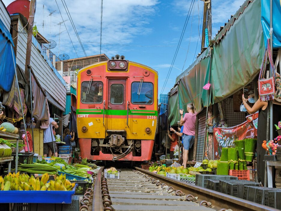 Das Bild zeigt den Mae-Klong-Markt südwestlich von Bangkok. Zu sehen ist eine orange Bahn, die in Schrittgeschwindigkeit frontal zum Betrachter durch die Engstelle des Mae-Klong-Marktes fährt. Links und rechts davon sind die Stände mit Obst und Gemüse, deren Planen vorübergehend eingezogen und deren die Auslagen zurückgerollt wurden. Die Bahn fährt sehr nah darüber hinweg. Link zur vergrößerten Darstellung des Bildes.