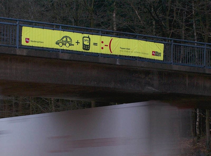 Zu sehen ist ein gelbes Transparent an einer Autobahnbrücke, das mit Piktogrammen auf die Niedersächsische Verkehrssicherheitskampagne „Tippen tötet“ hinweist.
