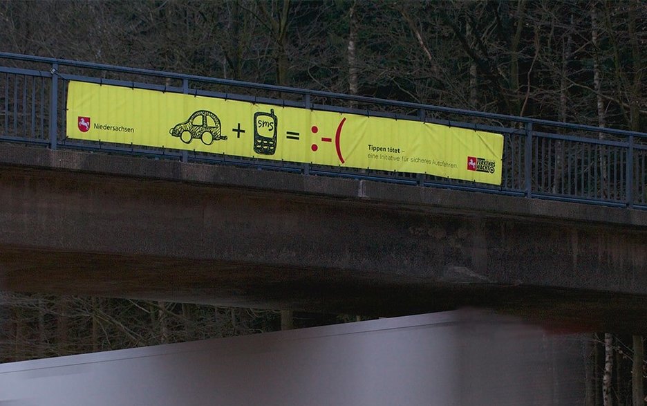Zu sehen ist ein gelbes Transparent an einer Autobahnbrücke, das mit Piktogrammen auf die Niedersächsische Verkehrssicherheitskampagne „Tippen tötet“ hinweist.