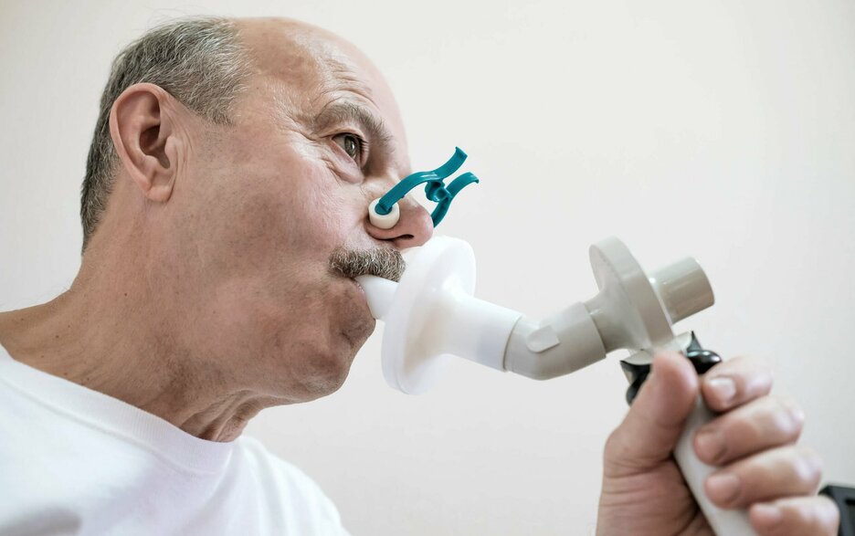 Auf dem Foto ist ein Mann beim Lungenfunktionstest zu sehen. Er trägt eine Nasenklammer und bläst in ein Messgerät hinein. Dieser Test gehört mit zur arbeitsmedizinischen Vorsorge bei staubintensiven Tätigkeiten. 