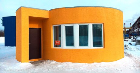 Das Bild zeigt ein orangefarbenes Betonhaus mit runden und eckigen Formen, das durch einen 3D-Drucker erschaffen wurde. Link zur vergrößerten Darstellung des Bildes.