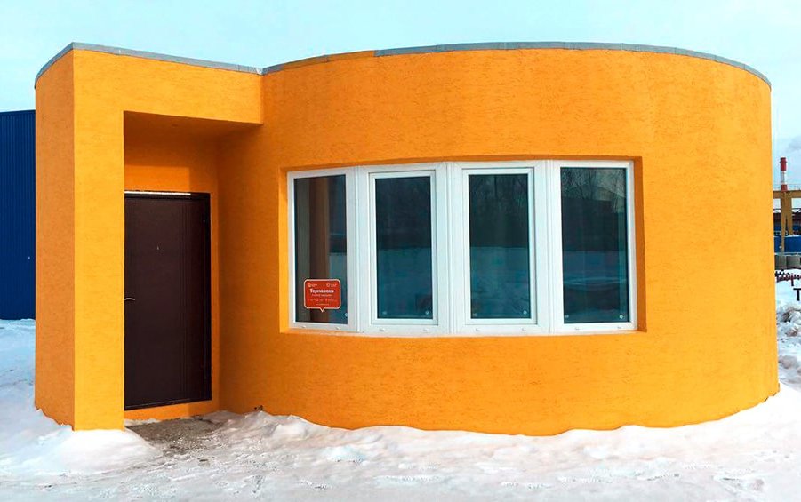 Das Bild zeigt ein orangefarbenes Betonhaus mit runden und eckigen Formen, das durch einen 3D-Drucker erschaffen wurde. Link zum Artikel.